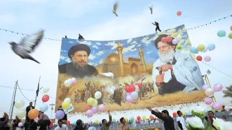 در این تصویر، تعدادی از مردم در حال برگزاری یک مراسم یا جشن هستند. در پس‌زمینه، یک دیوار بزرگ با تصویری از دو شخصیت سیاسی یا مذهبی به نمایش درآمده است. افراد در حال پرتاب بالن‌های رنگی و همچنین برخی گل‌ها به سمت هوا هستند. برخی از مردم در حال رقص و شادمانی دیده می‌شوند. پرنده‌هایی نیز در آسمان در حال پرواز هستند و فضای کلی تصویر روحیه مثبت و شادابی را به نمایش می‌گذارد.