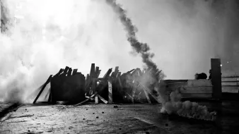 L'image montre une scène de tension, probablement associée à une manifestation ou à des troubles civils. On y voit des barricades faites de planches de bois, qui semblent être disposées sur une route. Une épaisse fumée s'élève dans l'air, créant une atmosphère sombre et chaotique. La monochromie de la photo ajoute à l'intensité de la scène, soulignant le contraste entre les ombres et la lumière. Des éléments comme des fumigènes ou des projectiles peuvent également être présents, suggérant une confrontation entre manifestants et forces de l'ordre. L'ensemble évoque une situation de conflit et de désobéissance civile.