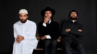 L'image montre trois hommes assis sur un banc, chacun portant un style vestimentaire distinct. À gauche, un homme en blanc, portant une tenue traditionnelle et un couvre-chef. Au centre, un homme habillé de manière formelle, avec un chapeau noir et une tenue sombre. À droite, un homme également en costume, avec une expression pensive. Le fond est noir, ce qui met en valeur les vêtements et les postures des sujets.