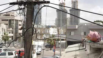 L'image montre une vue urbaine où plusieurs poteaux électriques sont entremêlés de fils. À l'arrière-plan, on peut apercevoir deux grands immeubles en construction. Au premier plan, il y a des bâtiments plus petits et des véhicules stationnés. On peut également voir des palmiers, ce qui indique un environnement peut-être chaud ou méditerranéen. L'ensemble donne une impression de ville en développement, avec un mélange de structures anciennes et modernes.