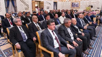 L'image montre un groupe de personnes assises, probablement lors d'une réunion ou d'une conférence. Ils semblent être des hommes d'affaires ou des responsables, vêtus de costumes formels. L'arrière-plan est orné de décorations élégantes, typiques d'une salle de conférence dans un cadre sophistiqué. Les participants regardent attentivement, suggérant un moment de discussion ou de présentation importante.
