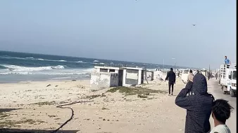 L'immagine mostra una scena costiera con il mare e delle onde in movimento. Si possono vedere alcune persone sulla spiaggia che osservano il mare, apparentemente interessate a qualcosa in cielo. Il paesaggio è caratterizzato da sabbia chiara e una scalinata che porta a una struttura, mentre in lontananza si vedono veicoli e persone che si muovono lungo la costa. Il cielo è sereno, con poche nuvole.