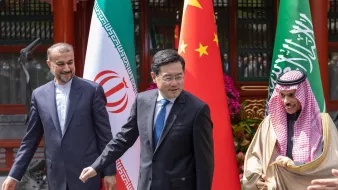 در این تصویر، سه نفر در حال ایستادن و گفتگو در یک محیط رسمی هستند. در سمت چپ، مردی با لباس رسمی و کت و شلوار تیره دیده می‌شود که با لبخند به سمت دیگران نگاه می‌کند. در وسط، مردی دیگری با کت و شلوار تیره و کراوات با رنگ آبی دیده می‌شود که به سمت راست می‌رود. در سمت راست، مردی با لباس سنتی عربی است که با لبخند به دوربین نگاه می‌کند. در پس‌زمینه، پرچم‌های ایران، چین و عربستان سعودی نمایش داده شده‌اند که نشان‌دهنده همکاری یا دیداری بین این کشورها است. فضای عمومی رسمی و دوستانه‌ای در تصویر وجود دارد.