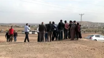 L'image montre un groupe de personnes rassemblées sur un terrain semi-aride. Ils semblent observer quelque chose au loin, peut-être un événement ou une scène particulière. L'arrière-plan est dégagé, avec des collines et des bâtiments visibles au loin. Il y a des véhicules stationnés, dont une voiture à droite. L'atmosphère suggère une interaction entre les gens, qui semblent concentrés sur ce qu'ils regardent. Des câbles électriques sont visibles, ajoutant à l'ambiance rurale de la scène.