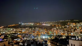 L'image montre une vue nocturne d'une ville illuminée. On peut voir de nombreux bâtiments agencés en terrasses, avec des lumières qui scintillent dans l'obscurité. À l'arrière-plan, des collines sont éclairées, et quelques structures sont visibles au sommet. L'atmosphère est tranquille, avec un ciel nocturne clair. Les antennes et structures en premier plan ajoutent une dimension urbaine à cette scène paisible.