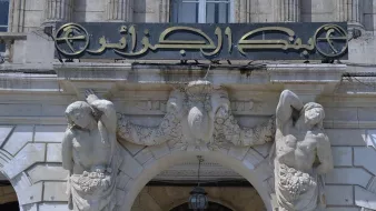 The image features a historical bank building with an ornate entrance. Above the doorway, there is a sign in Arabic that reads "بنك الجزائر" which translates to "Bank of Algeria." The architecture includes decorative elements, such as sculptures of two male figures on either side of the entrance. These figures appear to be holding or surrounded by garlands or fruit, adding to the classical decorative style of the building. The overall design is elaborate, reflecting a historical architectural style.