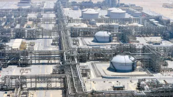 L'immagine mostra un impianto industriale, probabilmente una raffineria o un complesso di estrazione di prodotti petroliferi. Si possono vedere grandi serbatoi rotondi e una vasta rete di tubazioni che si snodano attraverso il sito. L'area circostante appare deserta, con una superficie di sabbia o terra arida. Sullo sfondo si intravedono strutture alte, che potrebbero essere torri di raffineria o impianti di trattamento. L'intero complesso è ben organizzato, con una disposizione sistematica delle tubazioni e dei serbatoi.