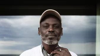 L'image montre un homme debout, portant une chemise blanche et un chapeau beige. Il est en train de nouer une écharpe autour de son cou. Son visage est marqué par l'âge, avec une barbe grise bien entretenue. Il porte également plusieurs bagues aux doigts et une montre en cuir. En arrière-plan, on peut voir une fenêtre qui donne sur une mer calme, sous un ciel nuageux. L'ensemble de l'image dégage une atmosphère tranquille et contemplative.