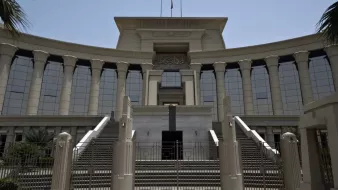 L'image montre un bâtiment imposant, probablement une institution judiciaire, avec une architecture moderne. La façade est ornée de colonnes majestueuses et de grandes fenêtres réfléchissantes, créant une impression de grandeur. Il y a des escaliers menant à l'entrée principale, qui semble être encadrée par des éléments décoratifs. Le ciel est dégagé, suggérant une journée ensoleillée. Des palmiers sont visibles sur les côtés, ajoutant une touche tropicale à l'environnement.