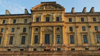 L'image montre un bâtiment imposant, probablement un palais ou un tribunal, éclairé par la lumière dorée du soleil couchant. La façade est ornée de détails architecturaux élaborés, avec des colonnes et des sculptures qui ajoutent à son majestueux style classique. Le nom "COUR DE CASSATION" est visible, indiquant qu'il s'agit d'un lieu judiciaire important. Le cadre montre un ciel clair, accentuant la beauté de l'architecture historique.