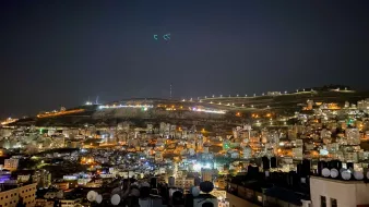 L'immagine mostra una vista notturna di una città, con luci brillanti che illuminano gli edifici. Sullo sfondo si possono vedere colline, anch'esse parzialmente illuminate. Gli edifici sono disposti su vari livelli, creando un effetto panoramico. La scena trasmette un'atmosfera vivace e dinamica, tipica di un centro urbano notturno. La presenza di antenne e strutture sul tetto di alcuni edifici suggerisce un'area residenziale o commerciale.