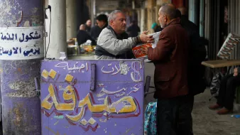 در تصویر، یک مرد در حال تبادل اطلاعات یا کالا با شخص دیگری است. دو مرد در جلوی یک دکه که بر روی آن نوشته‌هایی به زبان عربی و انگلیسی وجود دارد، ایستاده‌اند. دکه به نظر می‌آید که مربوط به تبادل کالا یا خدمات است. محیط اطراف شلوغ و بازاری به نظر می‌رسد که نشان‌دهنده زندگی روزمره در یک محل شهری است. رنگ‌های دکه روشن و نوشته‌ها واضح هستند.