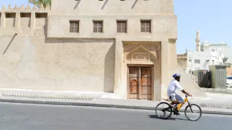 L'image montre un homme à vélo qui passe à côté d'un bâtiment au style architectural traditionnel. Ce bâtiment, de couleur sable, présente des murs texturés et des fenêtres ornées de motifs. On peut apercevoir une grande porte en bois sculpté, typique de l'architecture ancienne. En arrière-plan, on voit une mosquée avec un minaret, ce qui évoque une ambiance tranquille et culturelle dans une ville du Moyen-Orient. Le ciel est dégagé et ensoleillé.