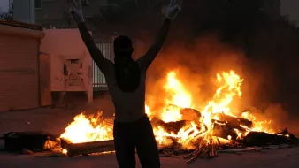 L'image montre une scène intense et dramatique. À l'avant-plan, une personne se tient avec les bras levés, formant un signe de victoire, tout en portant un masque qui couvre son visage. En arrière-plan, on peut voir des flammes vives qui s'élèvent, indiquant un feu, probablement une barricade ou des objets en feu. L'atmosphère est chargée d'émotion, évoquant un moment de protestation ou de turbulence. Les ombres des bâtiments environnants ajoutent à l'ambiance sombre de la scène.