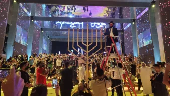 الصورة تظهر حدثًا احتفاليًا في مكان كبير. في المركز، يوجد هيكل على شكل منارة (المناورة)، مما يشير إلى الاحتفال بمناسبة معينة. يتم تنظيم الحضور حول الهيكل، وهناك أشخاص يحملون هواتفهم لتصوير الحدث. بعض الحضور يرتدون ملابس تقليدية، بينما يرتدي آخرون ملابس رسمية. هناك إضاءة ملونة وتفاصيل زينة جميلة في الأجواء، مما يضيف جوًا احتفاليًا حيويًا.