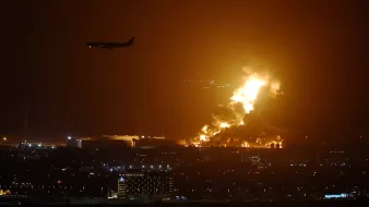 L'image montre un paysage nocturne éclairé par des flammes intenses, suggérant un incendie ou une explosion. On peut voir des colonnes de feu se levant dans le ciel, créant une ambiance dramatique. À l'arrière-plan, un avion est en train de survoler la scène, ce qui indique la proximité d'un aéroport. En bas, des lumières de la ville sont visibles, ajoutant un contraste entre l'urbanité et la catastrophe en cours.