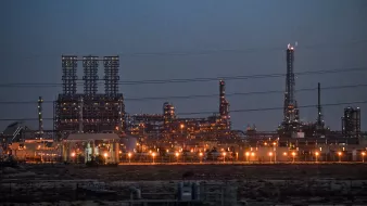 La imagen muestra una instalación industrial, probablemente una refinería o planta petroquímica, iluminada al anochecer. Se pueden ver numerosas estructuras metálicas altas, chimeneas y torres, con luces brillantes que resaltan en el entorno oscuro. Hay líneas eléctricas que cruzan la imagen y el paisaje es árido, lo que sugiere que la planta está ubicada en una zona desértica o poco vegetada. La atmósfera transmite una sensación de actividad industrial, con un enfoque en la tecnología y la producción.