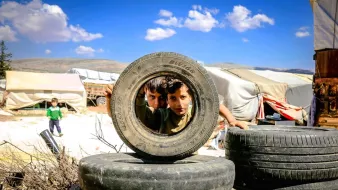 در این تصویر، دو کودک در حال نگاه کردن از میان یک لاستیک بزرگ هستند. لاستیک‌ها به صورت عمومی به عنوان بخشی از یک ساخت و ساز یا بازی در محوطه‌ای از فضای باز قرار داده شده‌اند. پس‌زمینه شامل چادرها و مناظر طبیعی است. آسمان آبی و ابرهای پراکنده در بالای سر دیده می‌شوند. فضای کلی تصویر حس کنجکاوی و بازی کودکانه را منتقل می‌کند.
