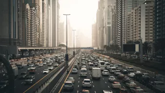 تظهر في الصورة مشهدًا حضريًا لمدينة مزدحمة، حيث تتواجد الكثير من السيارات المتوقفة في زحام مروري على عدة مسارات. تحيط بالمكان ناطحات السحاب العالية التي تدل على التطور العمراني في المنطقة. تبدو السماء مغشاة بضباب خفيف، مما يعطي انطباعًا عن أجواء المدينة في وقت متأخر من اليوم أو خلال الصباح الباكر. هناك مساحات من الأشجار على جوانب الطريق تضفي لمسة من الطبيعة وسط الحضر.