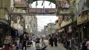 L'image montre une rue animée d'un quartier urbain. On peut y voir des bâtiments étroits et des fils électriques visibles. Des drapeaux et des affiches, probablement liés à un événement ou à une célébration, décorent l'entrée de la rue. Des personnes circulent, certaines à pied et d'autres à moto, ajoutant à l'activité de la scène. Le lieu semble être un marché ou un centre commercial, avec des étals vendant divers produits. L'atmosphère est vibrante et vivante.