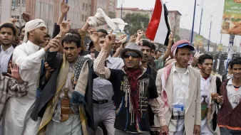 L'image montre un groupe de personnes en train de manifester dans une rue. Elles semblent exprimer des revendications, levant les bras et brandissant des drapeaux, notamment un drapeau rouge. Les manifestants portent des vêtements traditionnels yéménites, avec divers styles de vêtements. L'ambiance paraît énergique et engagée, comme une expression de protestation ou de soutien à une cause.