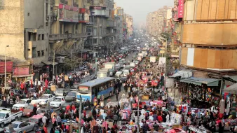 L'image montre une scène animée d'un marché urbain bondé, probablement dans une grande ville. On peut voir des foules de personnes se mêlant aux voitures dans une rue étroite. Les étals colorés sont remplis de divers produits, tels que des vêtements et des accessoires. Les bâtiments environnants semblent anciens et plusieurs niveaux sont visibles, avec des enseignes visibles. L'atmosphère paraît vivante et dynamique, typique des marchés populaires dans les grandes agglomérations.