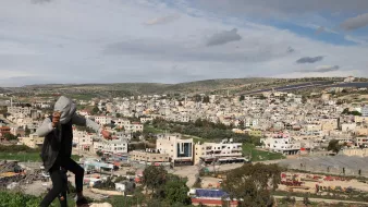 Nell'immagine si vede un giovane che si sta arrampicando su una roccia, con una città sullo sfondo. La città è composta da edifici di diversi piani, molti dei quali di colore chiaro. L'area è circondata da colline verdi e il cielo è parzialmente nuvoloso, suggerendo un clima variabile. Si possono notare anche alcune strutture agricole e aree di terreno aperto in primo piano.