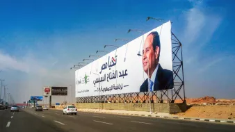 L'image montre un grand panneau publicitaire situé le long d'une route. Sur ce panneau, il y a une photo d'un homme en costume, accompagné d'un texte en arabe. La scène est prise dans un environnement urbain, avec une route et des véhicules en mouvement. Le ciel est clair et présente des teintes de bleu. Le panneau semble promouvoir une personnalité politique ou un événement lié à celle-ci.