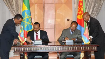 L'image montre une cérémonie de signature, probablement entre des représentants de l'Éthiopie et de l'Érythrée. Deux hommes, habillés en costume, signent des documents sur une table. À côté d'eux, des drapeaux éthiopien et érythréen sont visibles, ajoutant un élément symbolique à la scène. Les hommes semblent engagés dans un moment formel d'accord ou de collaboration.