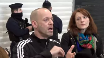 L'image montre un homme qui parle, visiblement en train d'expliquer quelque chose avec des gestes. Il porte un survêtement noir avec des rayures blanches. À côté de lui, une femme l'écoute attentivement, les cheveux bruns et portant un manteau noir. À l'arrière-plan, on peut voir des policiers en uniforme, certains portant des masques. L'ensemble suggère une situation de discussion ou d'intervention, possiblement liée à un événement impliquant les forces de l'ordre.