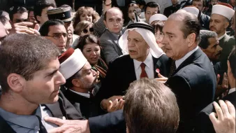 L'image montre une scène chargée d'émotion et de tension. On y voit plusieurs personnes rassemblées autour d'une figure centrale, visiblement un homme en costume. D'autres personnes, portant des vêtements traditionnels et des turbans, semblent interagir avec lui. L'atmosphère est dynamique, avec des visages concentrés et des gestes animés, ce qui suggère une discussion ou un événement important. Le mélange de styles vestimentaires indique une diversité culturelle parmi les personnes présentes, ce qui pourrait refléter un moment significatif sur le plan social ou politique.