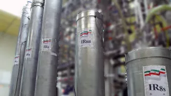 La imagen muestra varios cilindros metálicos dispuestos en un laboratorio o instalación industrial. Cada cilindro tiene una etiqueta que indica "IR8" o "IR8b", probablemente refiriéndose a modelos de centrifugas. En las etiquetas se puede ver una representación de la bandera de Irán. El fondo está desenfocado, pero se pueden observar más estructuras relacionadas con el equipo técnico, sugiriendo un ambiente relacionado con la investigación o la producción.