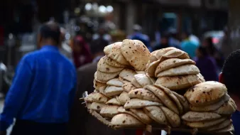 L'image montre un vendeur de pain dans une rue animée. Il porte une grande corbeille chargée d'un tas de pains ronds et dorés, souvent appelés "pita" ou "naan". En arrière-plan, on aperçoit des silhouettes de personnes se déplaçant dans la rue, créant une atmosphère vivante et dynamique. L'environnement semble plutôt urbain, avec des bâtiments et d'autres vendeurs visibles au loin.