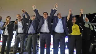 L'image montre un groupe de personnes sur une scène, levant les bras en l'air dans un geste de célébration ou de triomphe. Ils semblent être réunis pour un événement politique, probablement après l'annonce de résultats électoraux. Les participants portent des vêtements formels, et l'ambiance paraît festive et unie. En arrière-plan, on peut voir des écrans avec des informations, ce qui suggère que c'est une occasion importante.