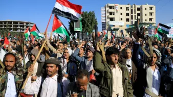 L'image montre une grande manifestation, probablement organisée par une foule, avec de nombreuses personnes levant des drapeaux, notamment des drapeaux aux couleurs de la Palestine. Les manifestants semblent déterminés et engagés, certains brandissant des banderoles. On peut voir des bâtiments en arrière-plan, suggérant un environnement urbain. L'atmosphère semble chargée d'énergie et de passion pour la cause.