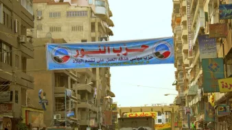 L'image montre une rue urbaine animée avec des bâtiments de plusieurs étages de chaque côté. Au-dessus de la route, il y a une grande banderole avec du texte en arabe. Cette banderole semble promouvoir un parti politique, qui pourrait être lié à des événements ou des activités locales. On aperçoit également des personnes marchant dans la rue, certaines portant des vêtements traditionnels. L'atmosphère de l'image évoque l'animation d'un marché ou d'un rassemblement communautaire.
