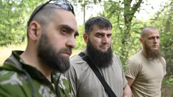 L'image montre trois hommes se tenant côte à côte dans une forêt. Tous trois portent des vêtements utilitaires, et chacun d'eux a une barbe. Leurs expressions sont sérieuses, et l'éclairage naturel met en valeur leur présence dans ce cadre boisé. On peut observer des arbres en arrière-plan, ce qui crée une ambiance naturelle.