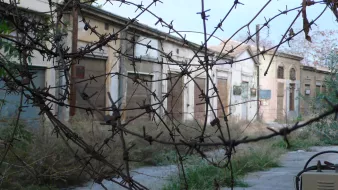 The image depicts an abandoned street scene, where several old buildings are visible with boarded-up windows and doors. The surroundings are overgrown with grass and weeds, suggesting neglect and disrepair. In the foreground, there are thorny branches or barbed wire, adding to the sense of desolation and possibly creating a feeling of confinement. The overall atmosphere feels eerie and forgotten, highlighting the passage of time in this once-active area.