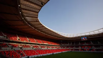 الصورة تظهر جزءًا من ملعب رياضي حديث. يتضح من التصميم الداخلي أن الملعب مزود بسقف عريض، مع صفوف من المقاعد الحمراء. الأجواء تبدو هادئة، حيث لا توجد جماهير في المدرجات، والسماء تظهر بلون أزرق فاتح. في أحد الجوانب، يمكن رؤية شاشة عرض كبيرة. الأرضية مغطاة بالعشب الأخضر، مما يضيف لمسة من الحيوية للمكان.