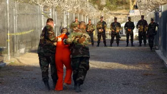 L'image montre un environnement militaire, probablement un centre de détention. On y voit des soldats en uniforme camouflé, certains tenant un individu vêtu d'une combinaison orange, qui semble être escorté. À l'arrière-plan, d'autres soldats sont alignés, surveillant la scène. Le tout se déroule dans un espace clôturé, avec des barbelés visibles, ce qui accentue l'atmosphère de sécurité et de contrôle. La lumière est naturelle, suggérant un moment en journée.
