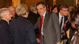 L'image montre un groupe de personnes réunies dans une salle, probablement lors d'un événement. Au centre, un homme en costume, avec une cravate à pois rouges, semble engager une conversation avec quelqu'un en lui serrant la main. D'autres personnes, en arrière-plan, observent ou participent à la discussion. L'ambiance semble conviviale, avec des sourires et des échanges entre les participants. Les murs sont décorés et ajoutent une touche d'élégance à la scène.