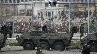 L'image montre une scène de tension, probablement dans une zone frontalière. On peut voir des véhicules militaires, dont un véhicule blindé, positionnés près d'une barrière. Des soldats sont en action, certains en uniforme et d'autres en tenue de combat, tandis qu'en arrière-plan, un grand groupe de personnes semble manifester ou se rassembler. La scène est marquée par une atmosphère de confrontation, avec des mesures de sécurité visibles.