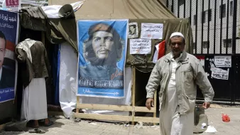 L'image montre un campement avec une tente, devant laquelle se trouve une affiche représentant un homme avec un béret, souvent associé à Che Guevara. À côté de l'affiche, on peut voir des affiches supplémentaires et une personne marchant, vêtue d'une tenue clair. L'ambiance semble refléter un contexte politique ou social, probablement avec des éléments de protestation ou de revendication.