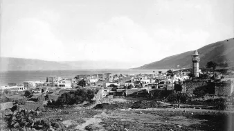 The image depicts a historical view of a coastal town near a body of water, likely a lake or sea, with a prominent lighthouse tower standing tall in the background. The landscape features a mix of stone buildings and ruins, suggesting a blend of architecture that may reflect the area's history. Surrounding the town are hills or mountains, adding depth to the scene. The overall mood is serene and nostalgic, capturing a moment from the past with a monochromatic or sepia tone.