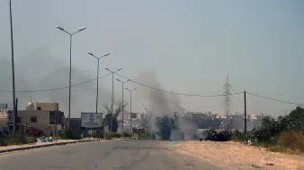L'image montre une scène de route apparemment désertée, avec quelques bâtiments sur les côtés. On peut apercevoir de la fumée s'élevant au loin, indiquant probablement un incendie ou une explosion. Des panneaux de signalisation sont visibles, ainsi que des lampadaires le long de la route. L'atmosphère semble chargée et la visibilité est atténuée par la fumée.