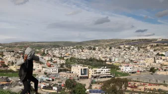 در این تصویر، فردی در حال بالا رفتن یا حرکت در مکانی کوهستانی دیده می‌شود. او لباس‌های غیررسمی به تن دارد و در پس‌زمینه، یک شهر بزرگ با ساختمان‌های مختلف و معماری متنوع دیده می‌شود. آسمان ابری است و مناظر طبیعی اطراف، مانند تپه‌ها و درختان، نیز در تصویر قابل مشاهده‌اند. این صحنه ترکیبی از زندگی شهری و طبیعت را نشان می‌دهد.