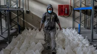 L'immagine mostra un uomo che cammina in un ambiente di magazzino. Indossa una felpa con cappuccio e una mascherina e tiene in mano due sacchetti bianchi. Intorno a lui ci sono numerosi sacchetti bianchi disposti ordinatamente sul pavimento. Le pareti del magazzino sono spoglie e ci sono scaffali metallici sullo sfondo. L'atmosfera sembra lavorativa e organizzata.
