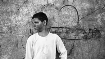 L'immagine mostra un ragazzo che sta in piedi di profilo, con un'espressione seria e pensierosa. Indossa una maglietta bianca e dei pantaloni chiari. Il suo viso appare segnato da segni di fango o polvere, il che suggerisce che potrebbe aver trascorso del tempo all'aperto o in un ambiente difficile. Dietro di lui, c'è un muro graffiato con disegni e segni, tra cui un cuore. L'atmosfera è in bianco e nero, conferendo un tono nostalgico e malinconico all'immagine.