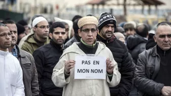 تظهر في الصورة مجموعة من الأشخاص يقفون في تجمع أو مظاهرة. في المقدمة، يوجد رجل يرتدي ملابس تقليدية ويقوم holding لافتة مكتوب عليها عبارة "PAS EN MON NOM" باللون الأبيض على خلفية بيضاء. يحيط به مجموعة من الرجال الذين يرتدون ملابس شتوية، ويبدو عليهم التوجه الجاد. تعكس الصورة أجواء من التعبير عن الرأي أو الاحتجاج.
