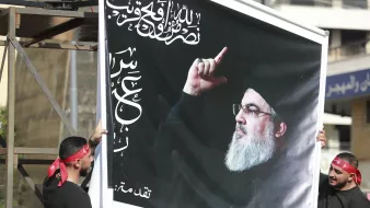 تصویر دو نفر را نشان می‌دهد که یک تابلو بزرگ را با تصویری از فردی در حال اشاره کردن به سمت بالا در دست دارند. پس‌زمینه تابلو سیاه است و بر روی آن متن‌هایی به زبان عربی دیده می‌شود. افراد حاضر در تصویر لباس‌های سیاه دارند و برخی از آن‌ها دستبندهای همرنگ بر روی سرشان دارند. این صحنه به نظر می‌رسد که در یک تجمع یا مراسم خاصی در حال برگزاری است.